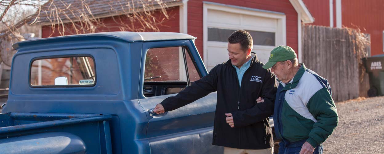 A caregiver opening the door of an old blue truck before helping his client into the passenger seat.