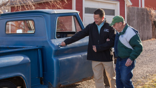 A caregiver opening the door of an old blue truck before helping his client into the passenger seat.