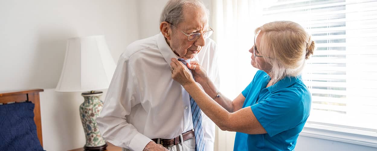 RAH-Website-Heroes-011720225-Desktop-1248x500 Female caregiver helping client put on his tie.