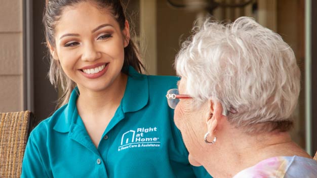 A female care worker smiling in the background. In the foreground is an elderly female patient smiling back.