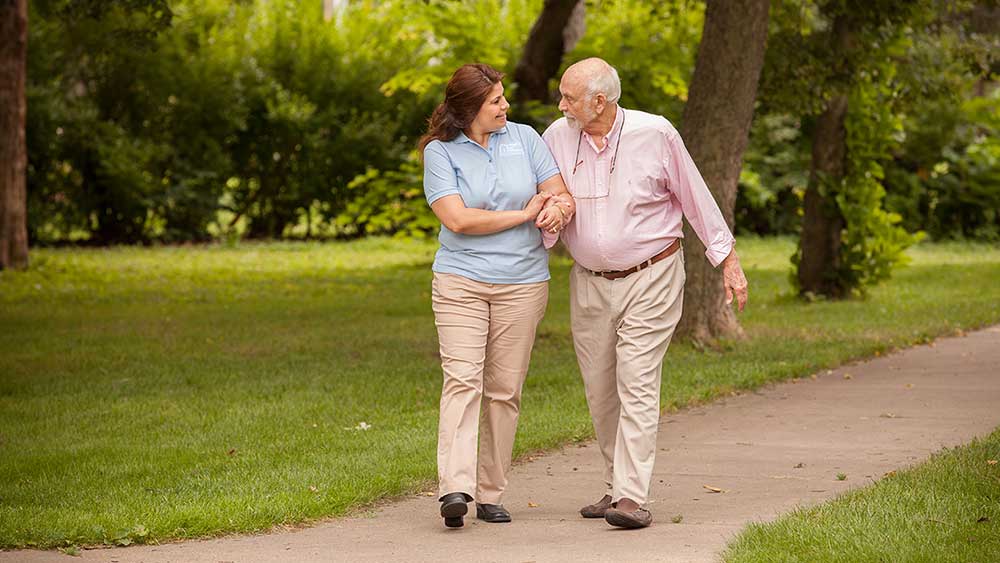 A female caregiver walking and talking with an elderly male patient.