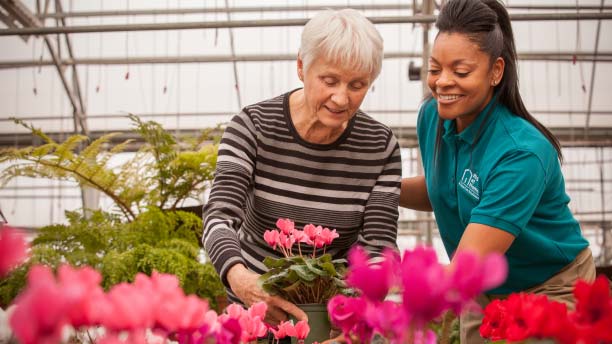A smiling female caregiver helping a smiling elderly woman pick out flowers in a market.
