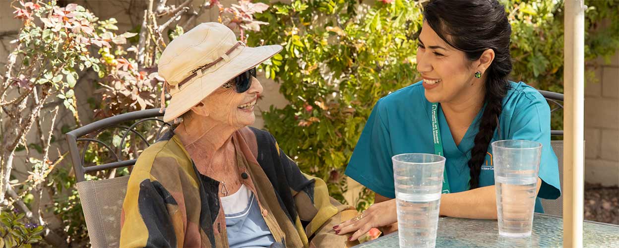 A female caregiver sits across from an elderly female patient outside with glasses of water. They both smile.