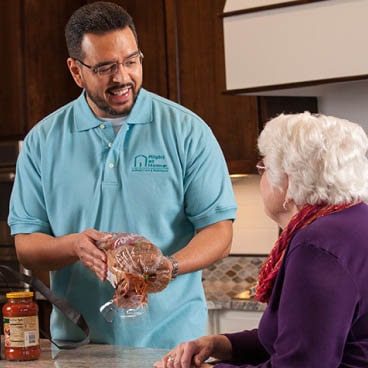 A male caregiver holds meal ingredients while talking and smiling with a female patient.