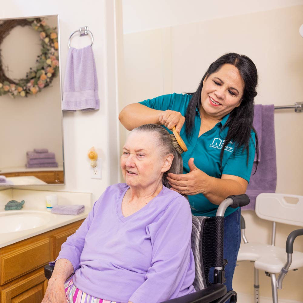 A female caregiver stands behind a female patient while brushing her hair.