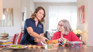 Female caregiver wearing mask serves plate of food to smiling elderly patient.