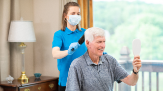 A female caregiver wearing a mask stands behind male patient while brushing his hair.