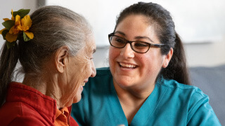 personal care hero image A female caregiver sits across from an elderly female patient, looking at her. They both smile.