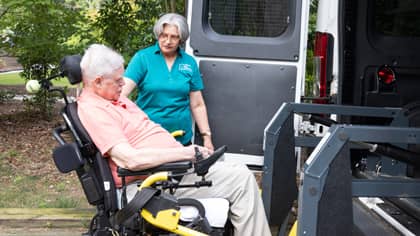 A female Right at Home caregiver helps an elderly disabled man in a wheelchair into a handy-capable van using a lift