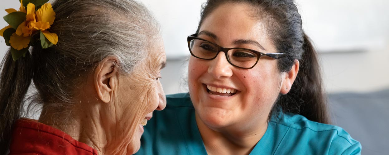 A female caregiver sits across from an elderly female patient, looking at her. They both smile.