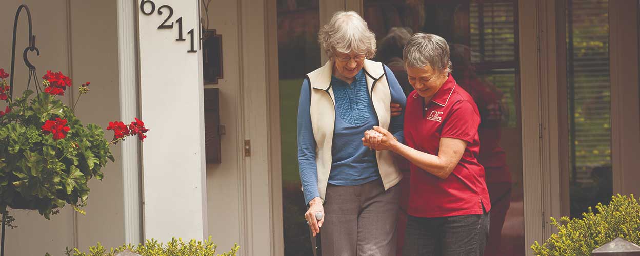 A female caregiver in red helps an elderly woman walk down stairs. They both smile.