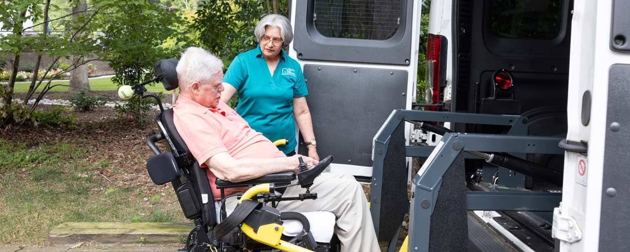 A female Right at Home caregiver helps an elderly disabled man in a wheelchair into a handy-capable van using a lift