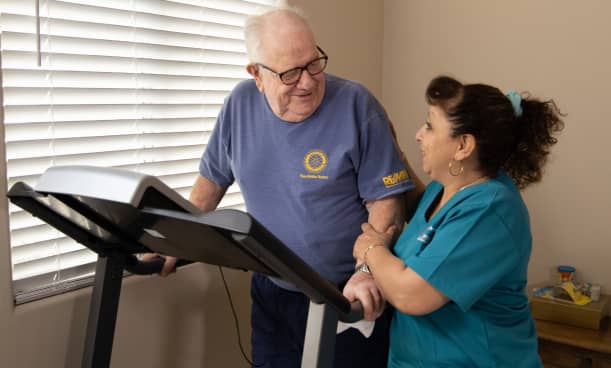 Female caregiver assisting elderly male patient on treadmill.